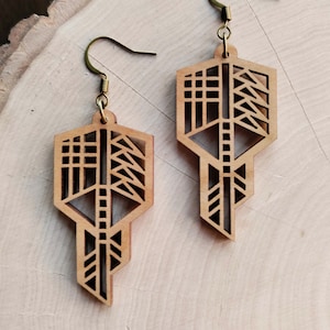 Puede incluir: Un par de pendientes geométricos de madera con acabado marrón claro. Los pendientes tienen forma de llave con intrincados diseños recortados y ganchos para las orejas de color latón. Se muestran sobre una superficie de madera.