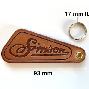May include: Brown leather keychain with the word "Simpson" embossed in a script font. The keychain has a metal ring with a 17 mm inner diameter and measures 3.9 inches by 3.7 inches.