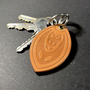 Può includere: Un portachiavi in pelle marrone con un portachiavi argentato e due chiavi. Il portachiavi ha una forma ovale con un disegno in rilievo di un toro stilizzato. Il portachiavi è lungo circa 7.6 cm.