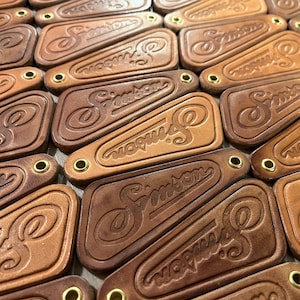 May include: A close-up of a pile of brown leather keychains with the word "Simpson" embossed on them. Each keychain has a brass ring.