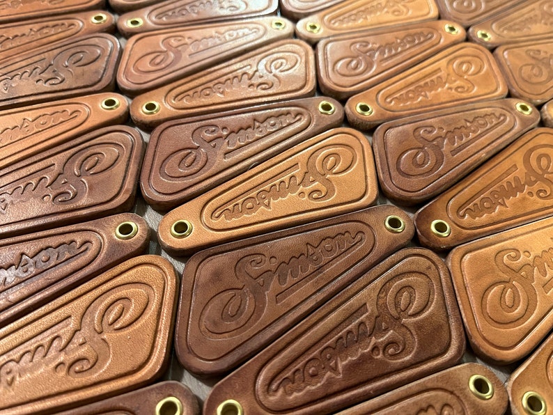 May include: A close-up of a pile of brown leather keychains with the word "Simpson" embossed on them. Each keychain has a brass ring.