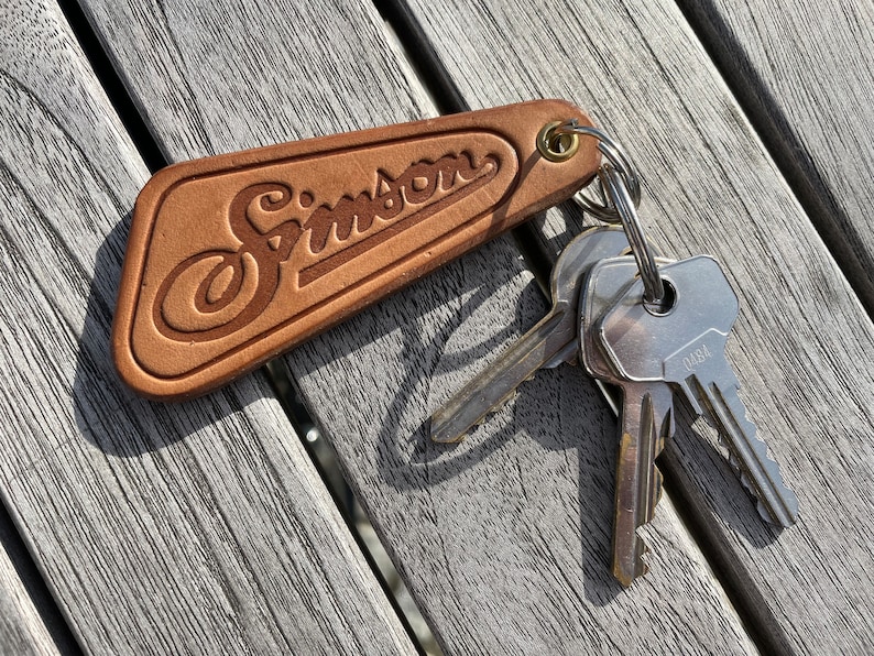 May include: A brown leather keychain with the word "Simson" embossed on it. The keychain has a silver key ring with three keys attached.