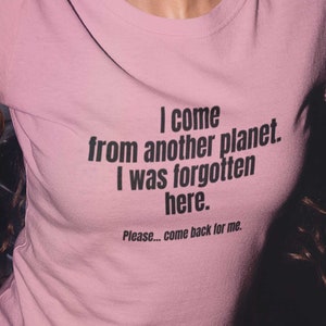 May include: A pink t-shirt with black text that reads "I come from another planet. I was forgotten here. Please... come back for me."