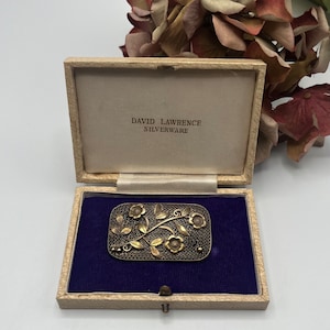 Op de afbeelding: Een zilveren en gouden bloemenbroche in een crèmekleurige doos met een blauwe fluwelen binnenkant. Op de deksel van de doos staat de tekst "DAVID LAWRENCE SILVERWARE" gedrukt.