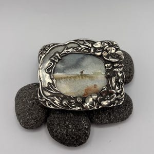 May include: A small, rectangular silver-toned frame with an art nouveau design, featuring a miniature watercolour painting of a seascape. The frame is adorned with floral and leaf motifs. The artwork rests on dark grey stones.