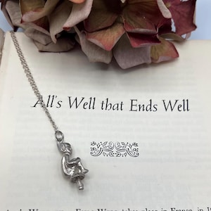 Puede incluir: Un marcapáginas plateado con una figura sentada, suspendida de una delicada cadena, descansa sobre un libro abierto. El título del libro, "All's Well that Ends Well", es visible. Flores secas en tonos burdeos y marrón en el fondo.