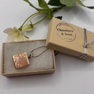 Pode incluir: Um pingente de relicário quadrado em ouro rosa numa delicada corrente de esferas, dentro de uma caixa de cartão bege com enchimento branco. A tampa tem um carimbo preto circular Chambers & Sons Antiques Vintage e um fio prateado.