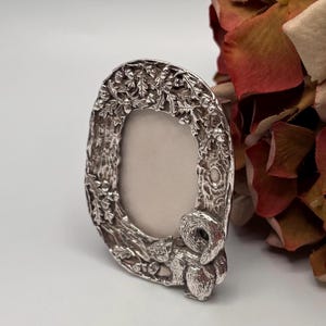 Peut inclure: Cadre photo ovale argenté avec un motif en relief détaillé. Le cadre représente un écureuil, des feuilles de chêne et des glands. Le cadre mesure environ 13 cm de haut et 10 cm de large, avec un espace ovale vide pour une photo.