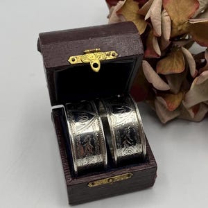 May include: Two silver napkin rings in a burgundy velvet box with a gold clasp. The rings are engraved with floral designs and the words "Grandmother" and "Grandfather".