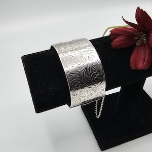 Peut inclure: Un large bracelet manchette argenté avec un motif floral gravé. Le bracelet est présenté sur un présentoir en velours noir, avec une petite chaîne attachée. Une fleur rouge foncé est en arrière-plan.