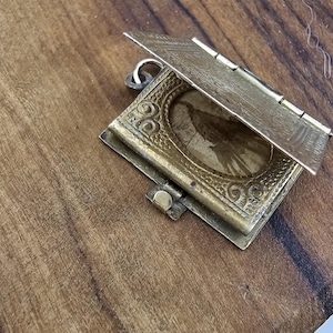 May include: A small, antique brass locket with an open lid, revealing a faded image. The locket is square with ornate detailing and a hinged top. It has a small clasp to secure the lid and a loop for a chain.