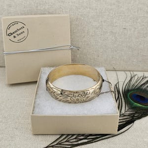 以下が含まれることがあります： 花と葉の彫刻が施されたゴールドカラーのヒンジ付きバングルブレスレット。白い内装のベージュ色の箱に展示されています。「Chambers & Sons Vintage」の文字が入ったお揃いの箱の隣にあります。背景には孔雀の羽があります。