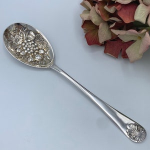 Puede incluir: Una cuchara de servir de plata con un diseño adornado de uvas y hojas. La cuchara tiene un mango largo y un cuenco redondeado.