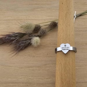 May include: A silver ring with a heart-shaped charm featuring the Gucci logo and the words "made in Italy". The ring is displayed on a wooden dowel, with dried flowers in the background.
