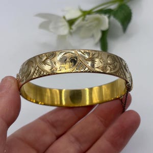 Könnte beinhalten: Ein goldfarbener Klapparmreif mit einem komplizierten Gravurmuster aus floralen Wirbelmotiven und schraffierten Texturen. Das Armband verfügt über einen sicheren Verschlussmechanismus und eine kleine Sicherheitskette an der Seite des Metallbandes.