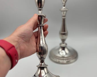 May include: Two silver candlesticks with ornate detailing. One is held by a hand with a red watch. The candlesticks have a wide base, a slender, ribbed body, and a top designed to hold a candle. The background is a light gray.