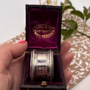 May include: A silver napkin ring with a decorative engraved pattern, housed in a dark brown rectangular box with a purple velvet interior. The inside lid features gold text reading HALL & CO SILVERSMITHS MANCHESTER.
