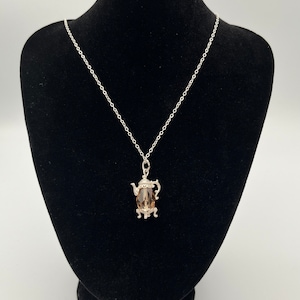 May include: A silver chain necklace with a brown crystal teapot pendant. The teapot has a detailed design and is set on three small feet.