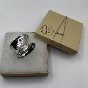 Peut inclure: Un rond de serviette en argent poli avec une forme concave évasée posé sur une boîte carrée beige avec un rembourrage en mousse blanche. Le couvercle porte un logo circulaire avec le texte Chambers & Sons Antiques Vintage et un élastique argenté.