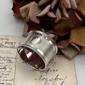 May include: A silver napkin ring with a textured design and a circular, smooth, raised center. The ring is lying on a piece of paper with handwritten text.