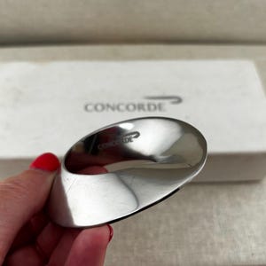 Könnte beinhalten: Ein polierter, silberfarbener Metall-Serviettenring mit einem glatten, gedrehten, ovalen Design. Die Oberfläche zeigt den eingravierten Text CONCORDE mit einem kleinen stilisierten Vogellogo. Er wird vor einer weißen rechteckigen Schachtel präsentiert.