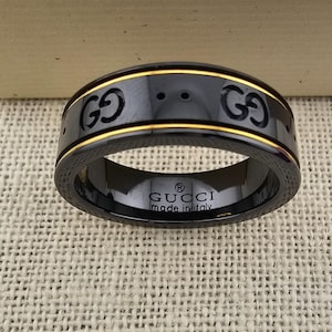 May include: Black ring with gold accents and the Gucci logo. The ring has a textured outer band and a gold stripe. The words "GUCCI made in italy" are printed on the inside of the ring.