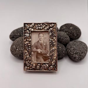 Pode incluir: Uma pequena moldura de fotografia antiga retangular com uma borda prateada ornamentada com folhas de carvalho e bolotas. A moldura contém uma fotografia em tons sépia de um homem de fato. A moldura está sobre pedras cinzentas escuras.