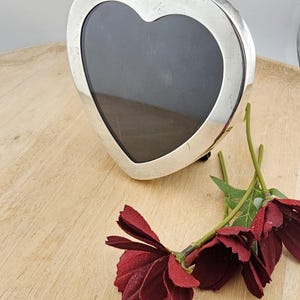 May include: A heart-shaped silver picture frame with a dark interior. The frame is on a light wooden surface, with burgundy-colored artificial flowers with green stems and leaves.