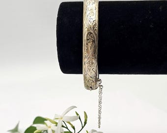 Könnte beinhalten: Ein silbernes Armreif-Armband mit einem eingravierten floralen Design. Das Armband wird auf einer schwarzen Samtoberfläche präsentiert. Eine feine Kette ist befestigt. Weiße Blumen und grüne Blätter sind im Vordergrund.