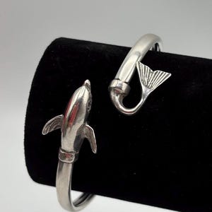 May include: A silver bracelet with a dolphin and a tail fin design. The bracelet is open and has a simple, elegant design.