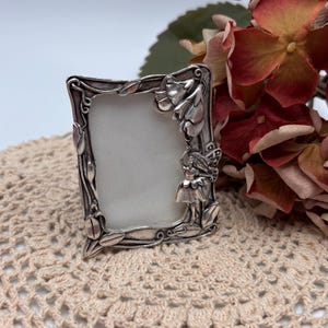 May include: A small, rectangular silver-toned picture frame with an ornate floral and fairy design. The frame has a blank white space for a photo and is set against a neutral background with flowers.