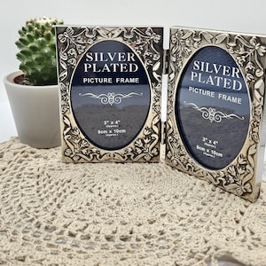 May include: A double silver-plated picture frame with an oval display area and embossed floral and ribbon detailing. The frame features text reading SILVER PLATED PICTURE FRAME and 3 x 4 inches, 8cm x 10cm. The hinged design allows for folding.