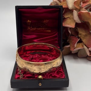 May include: A gold-coloured hinged bangle bracelet with an engraved floral design, presented in an open black box with a red velvet interior. The box displays the bracelet's details and a small chain.