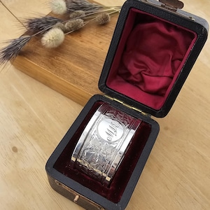 Puede incluir: Un brazalete de plata con patrones florales grabados e iniciales en un diseño circular. El brazalete se exhibe en una caja negra con interior de terciopelo rojo. La caja está abierta.