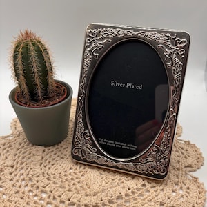 May include: A silver-plated rectangular photo frame with an oval centre opening and embossed ribbon and leaf detailing. The frame contains a black insert with the text Silver Plated and instructions to put the glass included in front.