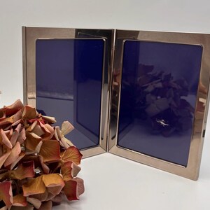 May include: A hinged, silver-toned metal photo frame with two panels, each displaying a deep blue interior. Dried, reddish-brown flowers are arranged in the foreground. The frame is reflective, with a polished finish, and is set against a white background.