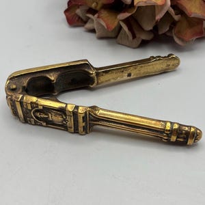 Peut inclure: Un casse-noix vintage en laiton avec un design orné. Le casse-noix est ouvert, révélant le mécanisme de cassage. Les poignées et le corps sont ornés de motifs décoratifs et d'un relief central. Le laiton a une patine chaude et vieillie.