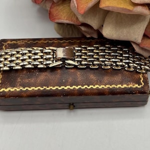 Può includere: Un braccialetto in metallo argentato con un design a rete poggia su un portagioie marrone vintage. La scatola ha un bordo dorato e una piccola chiusura. Un gruppo di fiori color pesca e gialli è sullo sfondo.