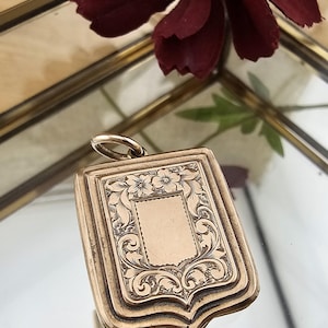 May include: A gold-colored, rectangular pendant with an ornate floral and scroll design. The pendant has a blank rectangular space in the center and a small loop at the top. The pendant is reflected in a mirrored surface.