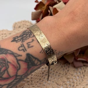 Peut inclure: Un bracelet jonc doré avec un motif floral gravé. Le bracelet est doté d'un fermoir et d'une chaîne de sécurité. Le motif est visible sur le devant du bracelet.