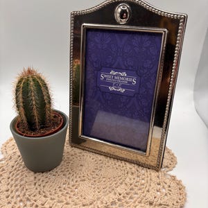 May include: A silver-toned metal photo frame with a beaded border and an arched top, featuring a purple insert with the text Sweet Memories Photo Frame 4 x 6 inches. Beside it sits a small green cactus in a matte gray pot.