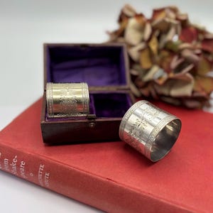 Puede incluir: Dos servilleteros plateados con grabados florales, uno dentro de una pequeña caja marrón con forro morado, el otro sobre un libro rojo. Los anillos tienen patrones detallados y un acabado pulido.