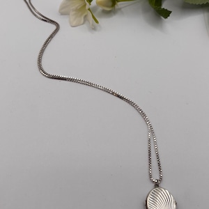 May include: A silver-toned oval locket pendant suspended from a delicate box chain. The locket features a textured surface with a series of radiating, curved lines that create a fan-like pattern across the front of the metal casing.