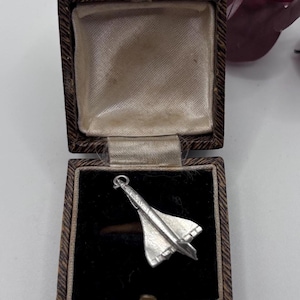 Könnte beinhalten: Ein silberfarbener Metallanhänger in Form eines Überschallflugzeugs mit spitzer Nase und Deltaflügeln. Der Anhänger befindet sich in einer kleinen, quadratischen Schmuckschatulle mit Klappdeckel, dunkelbrauner Texturoberfläche und schwarzem Samtfutter.