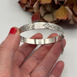 May include: A silver bangle bracelet with a decorative geometric pattern along the top edge. The bracelet has a hinged design and a safety chain. The bracelet is held in a hand with red nail polish.
