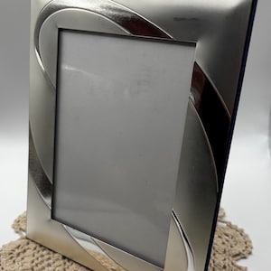May include: A rectangular silver-toned metal picture frame featuring a smooth, polished finish with curved, sweeping decorative accents along the border. The frame has a vertical orientation and a central rectangular opening for a photograph.