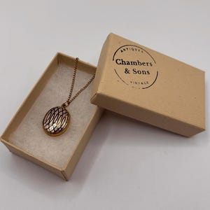 Puede incluir: Un collar con medallón dorado y diseño estampado, presentado en una caja de cartón abierta. El medallón tiene un colgante ovalado detallado. La caja lleva la inscripción "Chambers & Sons Vintage Antiques."