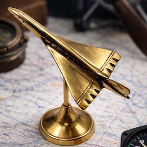 Könnte beinhalten: Ein goldfarbenes Metallflugzeugmodell, wahrscheinlich eine Concorde, wird auf einer Karte präsentiert. Das Modell ist auf einem runden Sockel montiert. Das Flugzeug hat detaillierte Flügel und ein schlankes Design. Ein Kompass und andere Navigationswerkzeuge sind im Hintergrund.