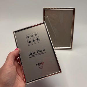May include: Two silver-plated brass photo frames. One frame is held by a hand, displaying the text "Silver Plated Brass Photo Frame" and the brand "SixTrees". The frames are designed for 4x6 inch photos.