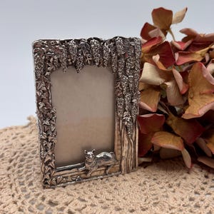 May include: A rectangular silver-toned picture frame with intricate floral and vine detailing. A sculpted cat rests at the base of the frame. The frame is empty, ready for a photograph. Dried flowers are in the background.